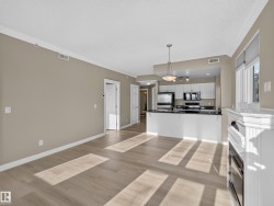 Open-concept living area with wood-finish flooring, crown molding, a built-in fireplace, and a kitchen featuring white cabinetry, dark countertops, and stainless steel appliances - 