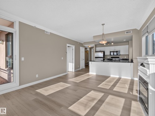 Open-concept living space featuring wood-finish flooring, a white trim fireplace, and a kitchen with white cabinetry, dark countertops, and stainless steel appliances - #1208 9939 109 St Nw, Edmonton, AB - Indoor Photo Showing Kitchen