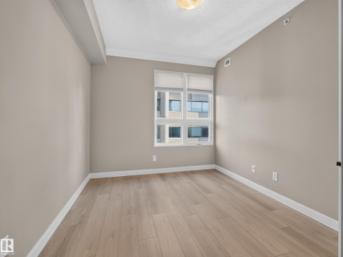 Neutral-toned interior featuring wood-finish flooring, a large window with roller shades, and crown molding - #1208 9939 109 St Nw, Edmonton, AB - Indoor Photo Showing Other Room