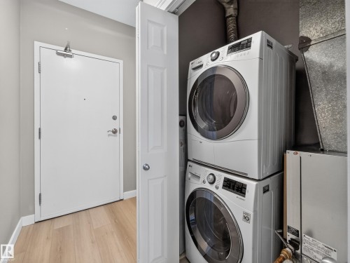 Dedicated laundry closet featuring a stacked washer and dryer, wood-finish flooring, white baseboards, and a solid core entry door - #1208 9939 109 St Nw, Edmonton, AB - Indoor Photo Showing Laundry Room