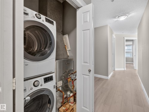 Dedicated laundry closet featuring a stacked washer and dryer, adjacent to a hallway with wood-finish flooring - #1208 9939 109 St Nw, Edmonton, AB - Indoor Photo Showing Laundry Room