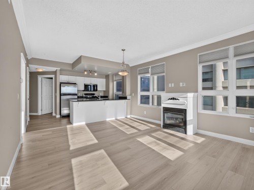 Open-concept living area featuring light wood-finish flooring, a contemporary fireplace with a decorative mantel, and a kitchen with white cabinetry and stainless steel appliances - #1208 9939 109 St Nw, Edmonton, AB - Indoor With Fireplace
