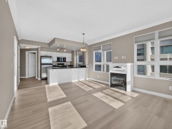 Open-concept living area featuring light wood-finish flooring, a contemporary fireplace with a decorative mantel, and a kitchen with white cabinetry and stainless steel appliances - 