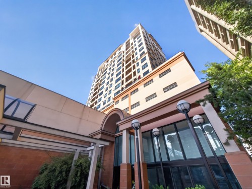 Modern high-rise exterior with a striking facade, featuring a prominent arched entrance, large storefront windows, contemporary exterior lighting fixtures, and a protective overhead walkway - #1208 9939 109 St Nw, Edmonton, AB - Outdoor