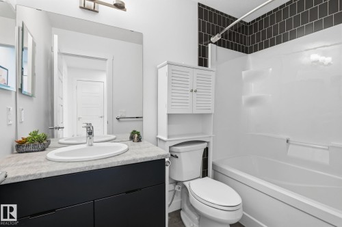 Bathroom featuring a dark vanity with integrated sink, a light-toned countertop, a frameless wall mirror, and a built-in tub with dark subway tile surround - 8282 Chappelle Way Sw, Edmonton, AB - Indoor Photo Showing Bathroom