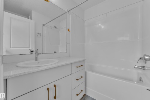Bathroom featuring a white vanity with an undermount sink, a light-toned countertop, brass hardware, and a white tub with a shower surround - #64 17832 78 St Nw Nw, Edmonton, AB - Indoor Photo Showing Bathroom