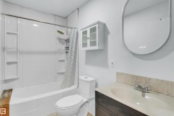 Bathroom featuring a white tub/shower combination, a wall-mounted storage cabinet, an oval mirror with integrated lighting, a vanity with a light-toned countertop, and a dark wood-finish vanity cabinet - 