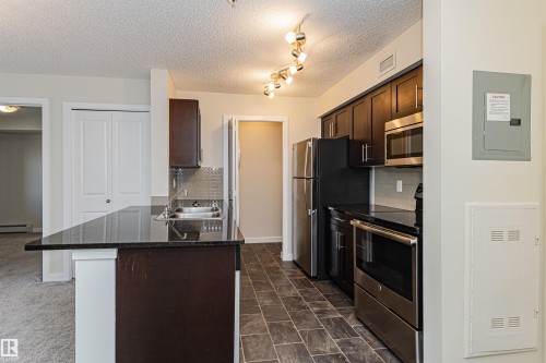 Kitchen featuring a breakfast bar with a black countertop, dark wood-finish cabinetry, stainless steel appliances, and tile flooring - #322 5816 Mullen Pl Nw, Edmonton, AB - Indoor Photo Showing Kitchen With Double Sink
