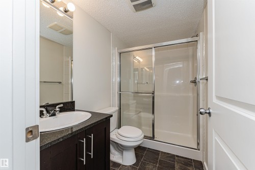 Bathroom featuring a dark wood-finish vanity with an integrated sink, a dark stone-finish countertop, and a shower with a sliding glass door - #322 5816 Mullen Pl Nw, Edmonton, AB - Indoor Photo Showing Bathroom
