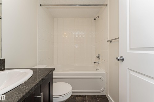 Bathroom featuring a built-in bathtub with white tile surround, dark wood-finish vanity with an integrated sink, and dark tile flooring - #322 5816 Mullen Pl Nw, Edmonton, AB - Indoor Photo Showing Bathroom