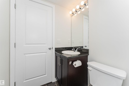 Bathroom featuring a dark wood-finish vanity, a black countertop, a white ceramic sink, and a mirrored vanity light fixture - #322 5816 Mullen Pl Nw, Edmonton, AB - Indoor Photo Showing Bathroom
