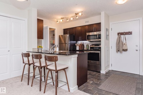 Kitchen featuring dark wood cabinetry, stainless steel appliances, and a black countertop island with seating - #322 5816 Mullen Pl Nw, Edmonton, AB - Indoor Photo Showing Kitchen