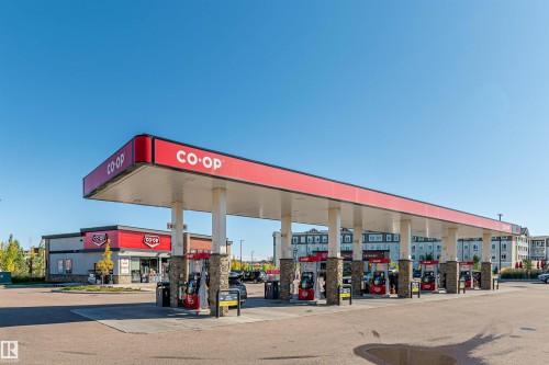 Retail fuel station featuring a prominent red canopy with white branding, multiple fuel pumps, stone-clad support pillars, and an accompanying convenience store building with coordinated signage - #322 5816 Mullen Pl Nw, Edmonton, AB - 