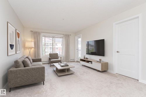 Spacious living area featuring light-toned carpeting, a large window with drapes, and a white interior door - #322 5816 Mullen Pl Nw, Edmonton, AB - Indoor Photo Showing Living Room