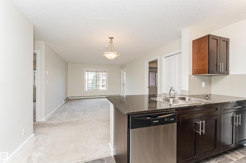 Kitchen island with a dark polished countertop, stainless steel sink, and integrated dishwasher - #322 5816 Mullen Pl Nw, Edmonton, AB - Indoor Photo Showing Kitchen With Double Sink