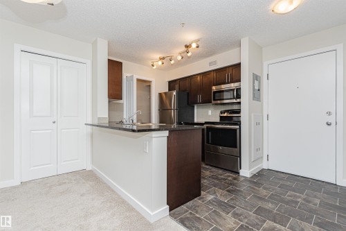 Modern kitchen featuring dark wood cabinetry, stainless steel appliances, a breakfast bar with a dark countertop, and a tile backsplash - #322 5816 Mullen Pl Nw, Edmonton, AB - Indoor Photo Showing Kitchen