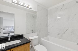 Contemporary bathroom featuring a white tile shower surround with gray veining, a white built-in bathtub, and a wood-finish vanity with a black countertop and integrated sink - 