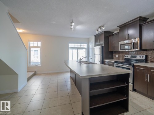 Open-concept kitchen featuring dark wood-finish cabinetry, stainless steel appliances, a mosaic tile backsplash, and a large island with an integrated sink and breakfast bar - #66 465 Hemingway Rd Nw, Edmonton, AB - Indoor Photo Showing Kitchen