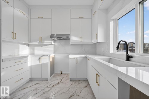 Modern kitchen featuring white cabinetry with gold-toned hardware, a stainless steel range hood, stone countertops, and a single-basin sink with a matte black faucet - 11 Harley Way, Spruce Grove, AB - Indoor Photo Showing Kitchen
