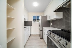 Modern kitchen featuring white cabinetry, light countertops, stainless steel appliances, a dark tile backsplash, and light wood-finish flooring - 