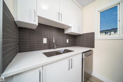 Modern kitchen space featuring white cabinetry with brushed nickel hardware, a stainless steel double basin sink with a gooseneck faucet, light-toned countertops, a dark gray tile backsplash, and a stainless steel dishwasher - 11432 139 Avenue Nw, Edmonton, AB - Indoor Photo Showing Kitchen With Double Sink