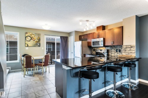 Open-concept kitchen and dining area featuring dark wood-finish cabinetry, stainless steel appliances, a multi-tone tile backsplash, and a breakfast bar with a dark countertop - 571 Orchards Boulevard Sw, Edmonton, AB - Indoor Photo Showing Kitchen