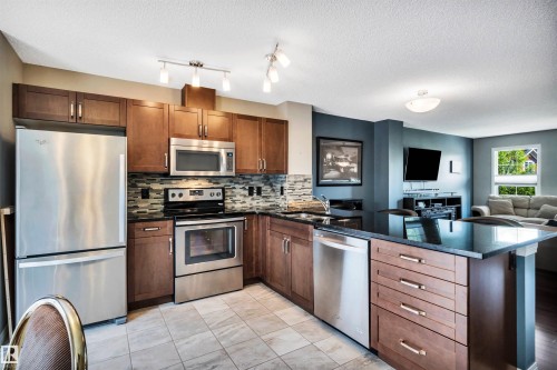 Kitchen featuring stainless steel appliances, dark wood cabinetry, black countertops, and a mosaic tile backsplash - 571 Orchards Boulevard Sw, Edmonton, AB - Indoor Photo Showing Kitchen