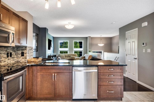 Kitchen island featuring a black countertop, integrated sink, stainless steel dishwasher, and rich wood cabinetry - 571 Orchards Boulevard Sw, Edmonton, AB - Indoor Photo Showing Kitchen With Double Sink