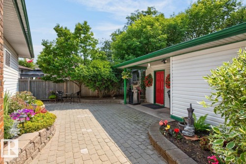 Paved stone patio featuring a vibrant garden bed with a stone retaining wall, mature trees, and a detached structure with white siding and a red entry door - 27 Milford Crescent, Sherwood Park, AB - Outdoor