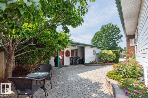 Expansive outdoor paved patio area featuring mature trees, landscaped garden beds, and white horizontal siding on the property structure - 27 Milford Crescent, Sherwood Park, AB - Outdoor With Deck Patio Veranda
