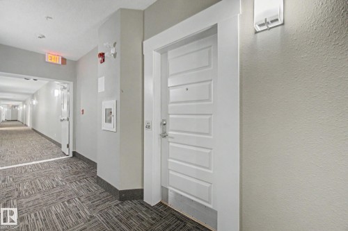 Corridor entrance featuring a panel door with contemporary hardware, light grey wall paint, and patterned carpet flooring - #435 1008 Rosenthal Bv Nw, Edmonton, AB - Indoor Photo Showing Other Room