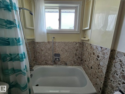 Bathtub with a patterned tile surround, white framed window, and a shower fixture with a flexible hose - 13103 63 Street Nw, Edmonton, AB - Indoor Photo Showing Bathroom