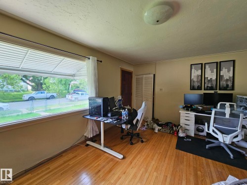 Spacious room featuring hardwood flooring, a large picture window with an exterior awning, a paneled door, bi-fold closet doors, and a ceiling-mounted light fixture - 13103 63 Street Nw, Edmonton, AB - Indoor Photo Showing Office