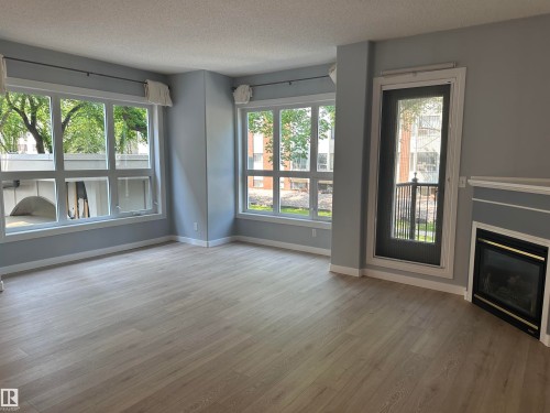 Spacious room featuring light wood-finish flooring, multiple large windows, a glass-paned exterior door, a built-in fireplace, and light gray wall tones - #203 11103 84 Av Nw, Edmonton, AB - Indoor Photo Showing Living Room With Fireplace