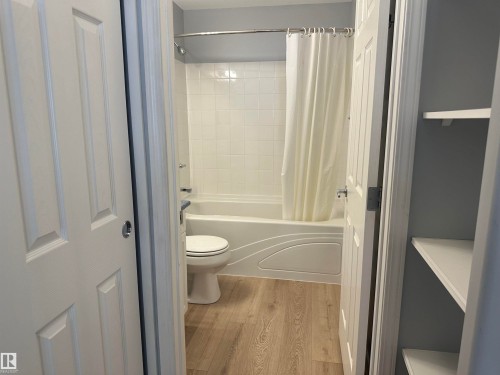 Bathroom featuring a white tub/shower combination with ceramic tile surround, a toilet, and wood-finish flooring - #203 11103 84 Av Nw, Edmonton, AB - Indoor Photo Showing Bathroom