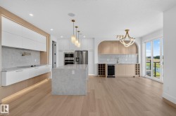 Modern kitchen and wet bar featuring wood-finish flooring, a large island with a stone-finish countertop, sleek white cabinetry, and stainless steel appliances - 