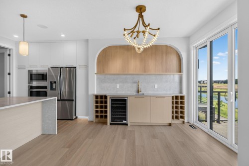 Open-concept kitchen and entertaining space featuring wood-finish flooring, a built-in wet bar with a beverage cooler, a stainless steel refrigerator, and sleek white cabinetry - 8791 Mayday Ln Sw Sw, Edmonton, AB - Indoor Photo Showing Kitchen With Upgraded Kitchen