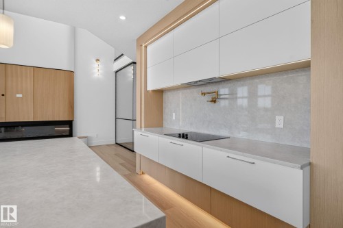 Contemporary kitchen featuring white flat-panel cabinetry, light wood-finish accents, integrated cooktop, stone countertops, and a gray stone backsplash - 8791 Mayday Ln Sw Sw, Edmonton, AB - Indoor Photo Showing Kitchen