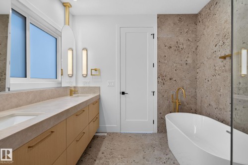 Contemporary bathroom featuring a freestanding soaking tub with a floor-mounted gold-tone faucet, a double vanity with light wood-finish cabinetry, integrated sinks, and a stone-look countertop - 8791 Mayday Ln Sw Sw, Edmonton, AB - Indoor Photo Showing Bathroom