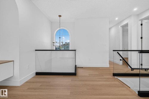 Spacious hallway featuring light wood-finish flooring, a modern glass and black metal railing, an arched window, a contemporary chandelier, and recessed lighting - 8791 Mayday Ln Sw Sw, Edmonton, AB - Indoor Photo Showing Other Room