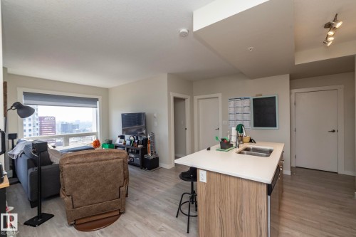 Open-concept living space featuring wood-finish flooring, a kitchen island with a double stainless steel sink and light-colored countertop, and a large window with city buildings visible - #2402 10410 102 Av Nw, Edmonton, AB - Indoor