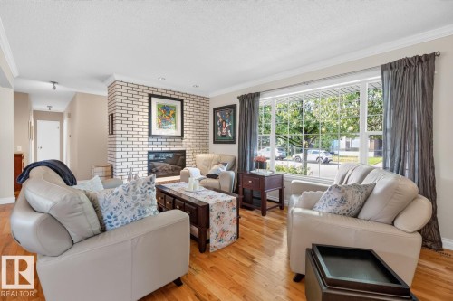 Living area featuring wood-finish flooring, a white brick fireplace with a black insert, and a large front-facing window - 4631 113A Street Nw, Edmonton, AB - Indoor Photo Showing Living Room With Fireplace