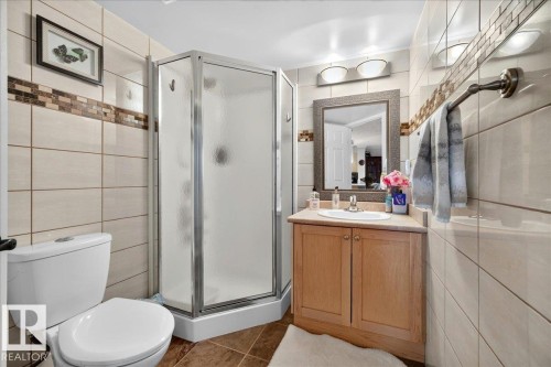 Bathroom featuring a corner shower enclosure with frosted glass, a light wood-finish vanity with a single sink, a framed mirror, and warm-toned wall tiling with a decorative mosaic accent band - #306 13111 140 Av Nw, Edmonton, AB - Indoor Photo Showing Bathroom