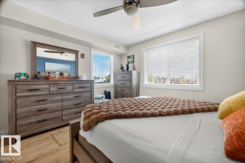 Bedroom with light wood-finish flooring, two large windows with horizontal blinds, and a ceiling fan - #306 13111 140 Av Nw, Edmonton, AB - Indoor Photo Showing Bedroom