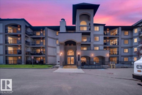 Multi-story residential building featuring an arched main entrance, multiple balconies with glass railings, a central tower element with arched windows, and a paved parking area - #306 13111 140 Av Nw, Edmonton, AB - Outdoor With Balcony With Facade