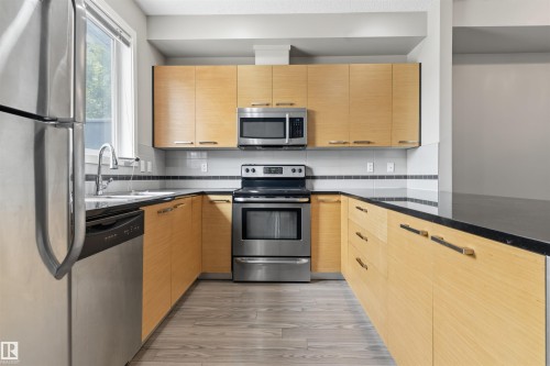 Open-concept kitchen featuring light wood-finish cabinetry, black countertops, stainless steel appliances, and wood-finish flooring - #62 675 Watt Bv Sw, Edmonton, AB - Indoor Photo Showing Kitchen
