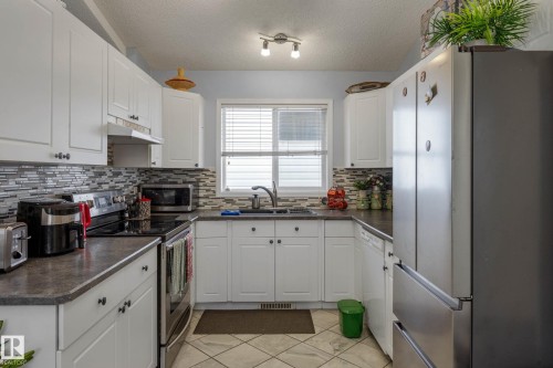 U-shaped kitchen featuring white cabinetry, dark countertops, a mosaic tile backsplash, and a stainless steel refrigerator - 9113 166 Avenue Nw, Edmonton, AB - Indoor Photo Showing Kitchen With Double Sink
