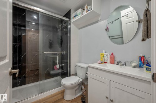 Bathroom featuring a glass-enclosed shower with black marble-effect wall tiling, a white vanity with integrated sink, wood-finish flooring, a wall-mounted shelving unit, and a circular mirror - 9113 166 Avenue Nw, Edmonton, AB - Indoor Photo Showing Bathroom