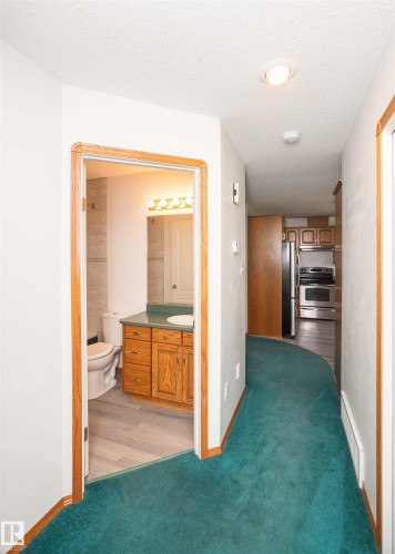 Hallway with teal carpeting and light wood trim, featuring an open doorway to a bathroom with wood-finish flooring, a white toilet, and a light wood vanity with a countertop and sink - 5116 51 Avenue, Pickardville, AB - Indoor Photo Showing Other Room