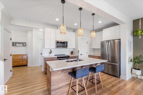 Contemporary kitchen featuring a large center island with integrated double sink, stainless steel appliances, white cabinetry, and wood-finish flooring - 6875 Evans Wynd Nw, Edmonton, AB - Indoor Photo Showing Kitchen With Double Sink With Upgraded Kitchen